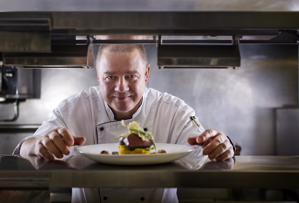Chef Jorg Penneke looks through the pass with a meat dish in the foreground