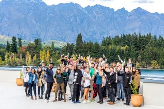 A group of hospitality workers raise wine glasses against the backdrop of Lake Wakatipu and The Remarkables