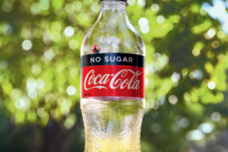 Empty coke bottle against nature background with "I'M NOW MADE FROM 100% RECYCLED PLASTIC" text