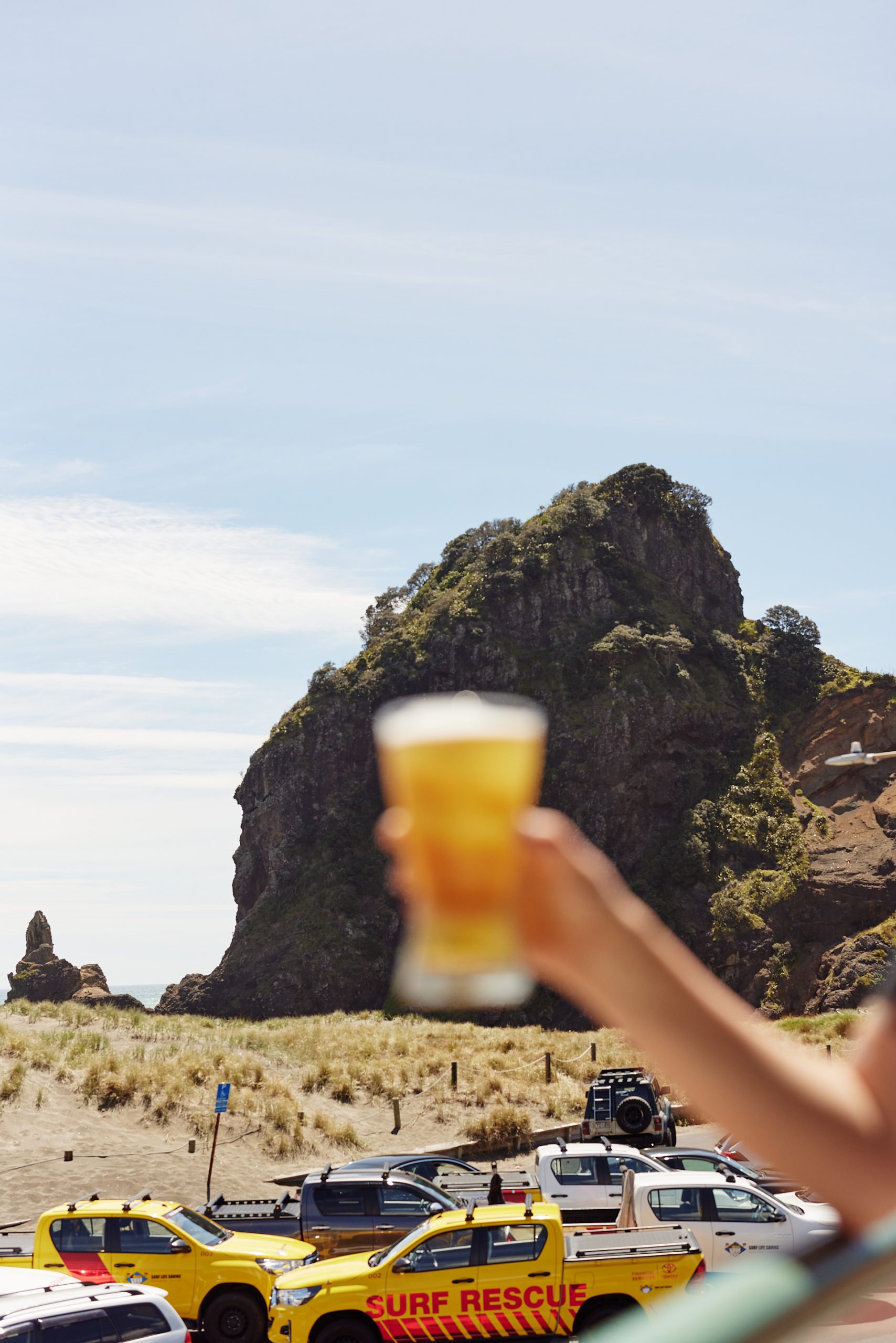 a beer held up against the view of the rock and sand with cars parked out the front