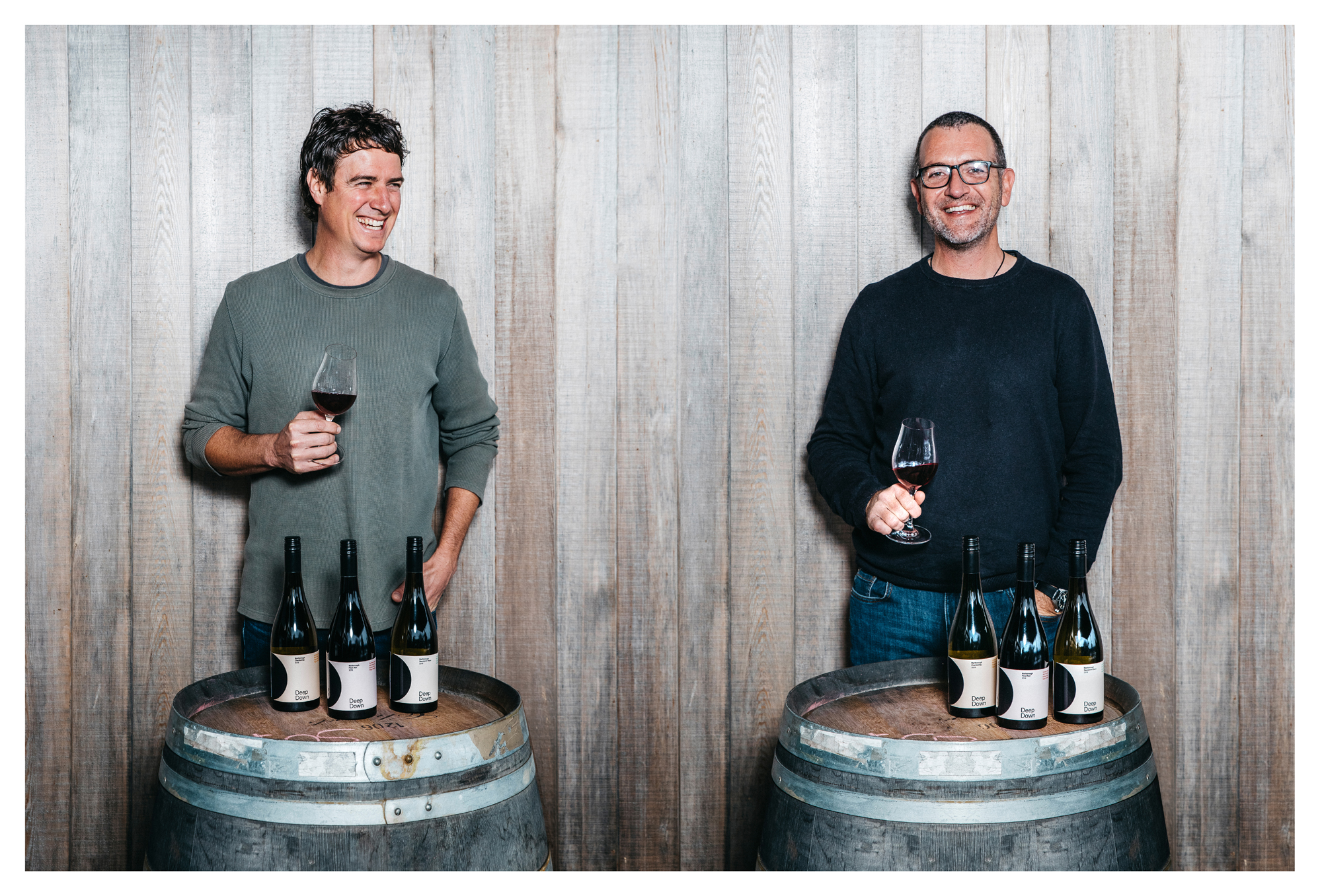 two men behind wine barrells with wine bottles on top