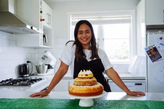 woman baker Jodeci Nolan standing in kitchen behind a three-tiered cheesecake in a kitchen